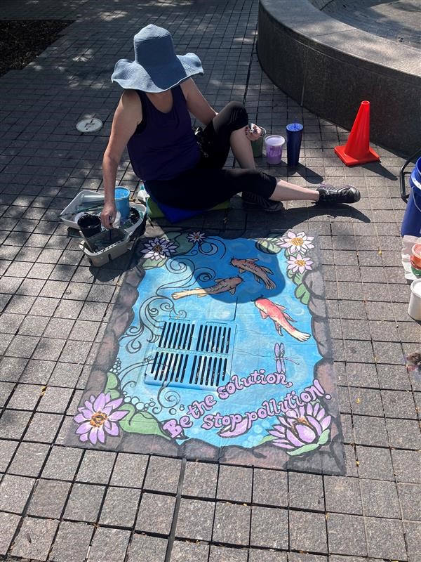 A sidewalk chalk mural depicting a koi pond with the words 'Be the solution, stop pollution!' The artist is sitting above the mural on the ground.